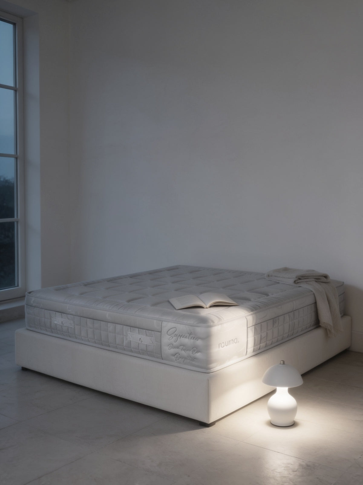 Signature-Matratze am Abend, am Boden leuchtet eine kleine Lampe, auf der Matratze liegt ein offenes Buch